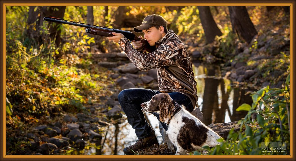 Young man posed outdoors by stream with hunting gun and dog