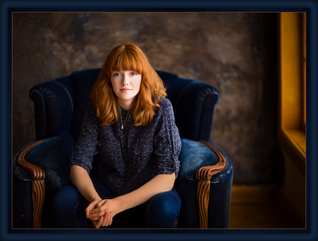 Senior portrait seated in navy velvet chair with rustic brown backdrop and natural window light