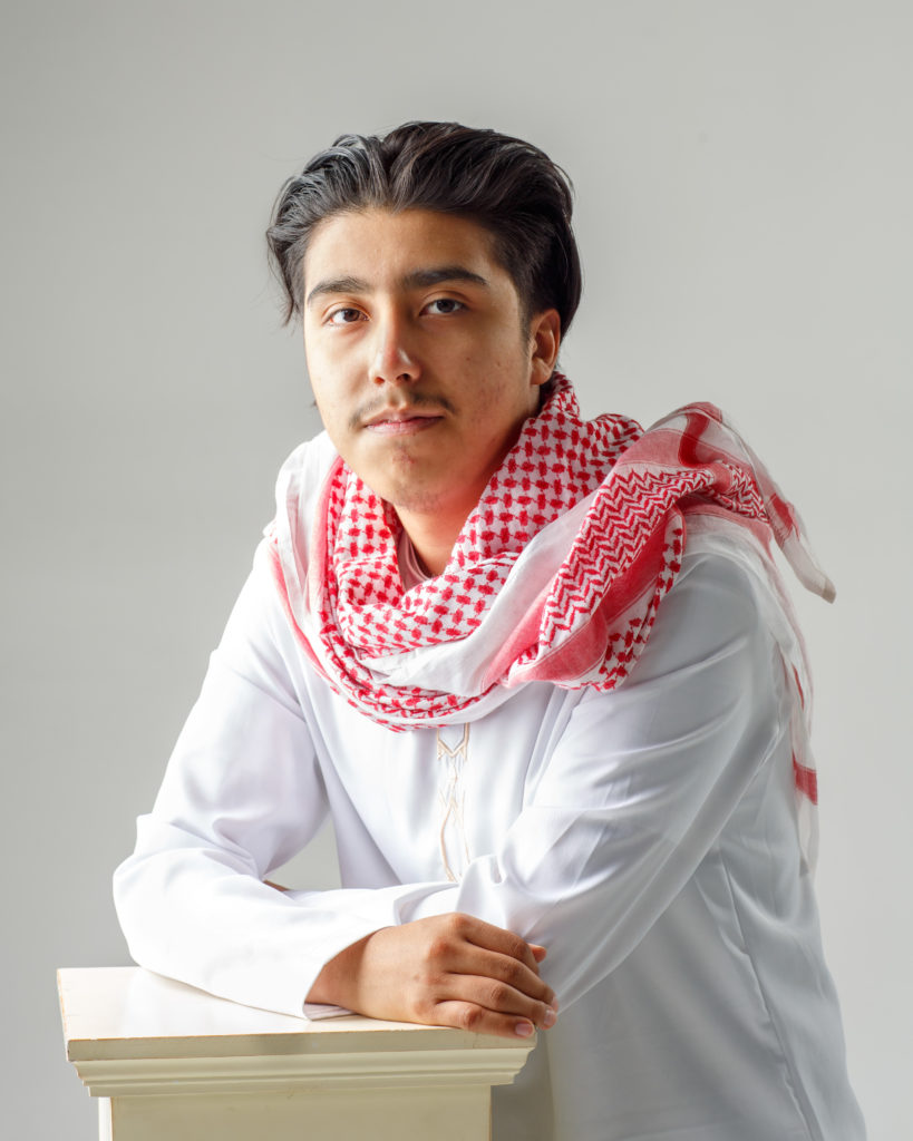Senior portrait in traditional white thobe and red keffiyeh, posed with arms crossed on white pedestal in studio setting